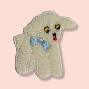 May include: A white felt lamb applique with a blue bow and yellow eyes. The lamb has a red tongue sticking out.