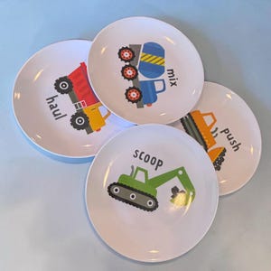 May include: A set of four white round plates featuring colorful construction vehicle illustrations. Each plate displays a specific vehicle with its corresponding action word printed in black: a red dump truck labeled haul, a blue mixer labeled mix, a green excavator labeled scoop, and an orange bulldozer labeled push.