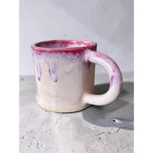 May include: A ceramic mug with a cylindrical body and a thick, curved handle. The mug features a light beige base color with a vibrant magenta glaze dripping from the rim down the sides, creating a textured, layered appearance on the surface.