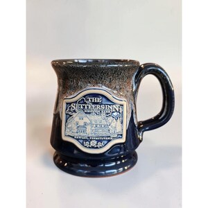 May include: A dark blue ceramic mug with a flared rim and a sturdy handle. The front features a cream-colored embossed emblem depicting a building with the text THE SETTLERS INN BINGHAM PARK HAWLEY, PENNSYLVANIA. The rim has a mottled brown glaze.