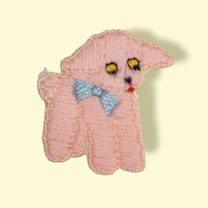 May include: A pink felt lamb with a blue bow tie. The lamb has embroidered eyes and a red mouth.