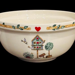 May include: Cream-colored ceramic mixing bowl with a decorative rim featuring a vine and leaf pattern. The bowl has a painted birdhouse scene with birds, a heart, and a small tree. The bowl is set against a black background.