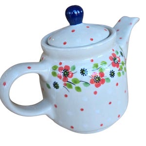 May include: A white ceramic teapot with a blue knob on the lid. The teapot is decorated with red flowers, green leaves, and red polka dots. The handle and spout are curved.