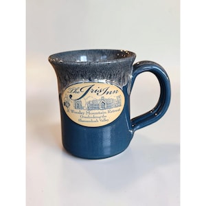 May include: A blue ceramic mug with a dark grey drip glaze at the rim. An oval cream-colored label on the front features the text The Iris Inn, A Woodsy Mountain Retreat, Overlooking the Shenandoah Valley, and an illustration of a building.