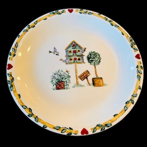 May include: A round, white decorative plate with a yellow border featuring green leaves and red hearts. The center of the plate has a painted scene of a birdhouse, birds, flowers, and trees. The plate is perfect for serving food or as a decorative piece.