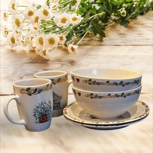 May include: A white ceramic dish set with floral and heart designs. The set includes two mugs, two bowls, and a saucer. The mugs and bowls have a decorative border with leaves and a wooden pattern. White flowers are in the background.