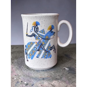 May include: A cream-colored ceramic mug with a speckled finish and a crackle glaze texture. The mug features a graphic illustration of a knight in silver armor riding a horse, both adorned in blue and yellow heraldic patterns with a sword.