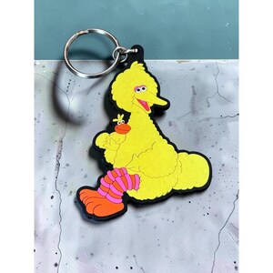 May include: A keychain featuring a yellow bird character with a long orange beak, pink and orange striped leg warmers, and large orange feet. The character is holding a small yellow duckling with an orange beak. The design has a black outline.