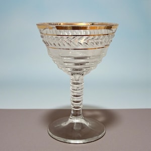 May include: A clear glass coupe with a gold rim and decorative leaf pattern. The stem is ribbed, and the base is round. The glass is designed for serving cocktails or champagne. The coupe is approximately 6 inches tall.