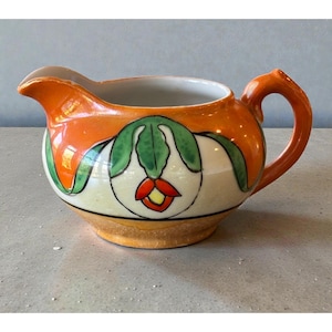 May include: A ceramic creamer with an orange exterior and a white interior. The creamer features a green and red floral design. The handle and spout are orange. The creamer sits on a light-colored surface.