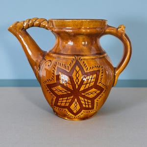 May include: A brown ceramic pitcher with a spout and handle. The pitcher features a large, star-shaped design in a darker brown color. The handle has a rope-like texture, and the spout is curved. The pitcher is likely used for pouring liquids.