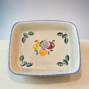 Könnte beinhalten: Quadratische Servierschale aus Keramik in Creme mit blauem Rand. Die Schale ist mit einem gemalten Fruchtdesign in der Mitte verziert, das eine gelbe Birne, eine lila Pflaume, eine rote Pflaume und eine orangefarbene Frucht zeigt. Grüne Blätter sind ebenfalls auf der Schale gemalt.