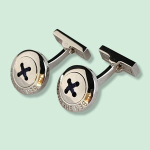 以下が含まれることがあります： ボタンのデザインが施されたシルバーのカフスボタンのペア。ボタンは黒で、中央に白いXがあります。カフスボタンには「BEN SHERMAN」という文字が刻まれています。