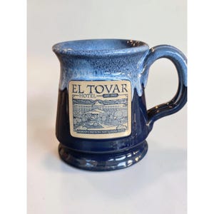 May include: A dark blue ceramic mug with a light blue drip glaze at the rim. A rectangular cream-colored label features a line drawing of the El Tovar Hotel with the text EL TOVAR HOTEL EST. 1905 GRAND CANYON NATIONAL PARK in dark blue ink.