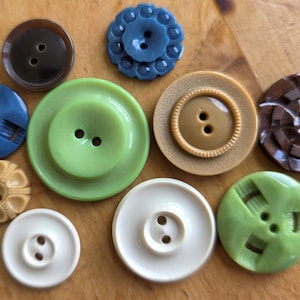 May include: A collection of various buttons in shades of brown, blue, green, and cream. The buttons vary in size, shape, and texture, including round, flower-shaped, and textured designs. Some have two holes, while others have four.