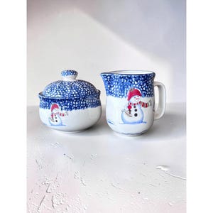 May include: A ceramic sugar bowl with a lid and a matching creamer pitcher, both featuring a white base, a blue speckled pattern at the top, and a hand-painted snowman wearing a red hat and scarf on the front of each piece.