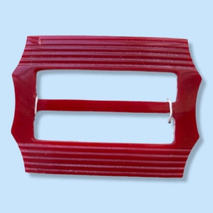 May include: A rectangular, red buckle with a ridged surface. The buckle has a central bar held in place by a silver-coloured metal piece. The edges of the buckle are shaped with decorative points.
