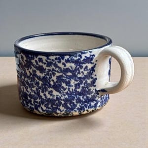May include: A handmade ceramic mug with a speckled blue and white pattern. The mug has a rounded shape, a sturdy handle, and a dark blue rim. The interior is white, and the mug appears to be well-crafted for everyday use.