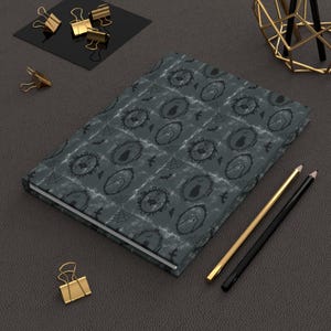 May include: A dark grey notebook featuring a repeating pattern of ornate oval frames containing silhouettes of ghosts, cats, and spiders, interspersed with small bats and spiderwebs, accompanied by two pencils and several gold metal binder clips.