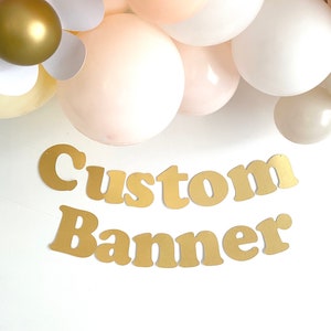 May include: Gold custom banner with the words "Custom Banner" in large, bold letters. The banner is hanging from a string of balloons in shades of white and beige.