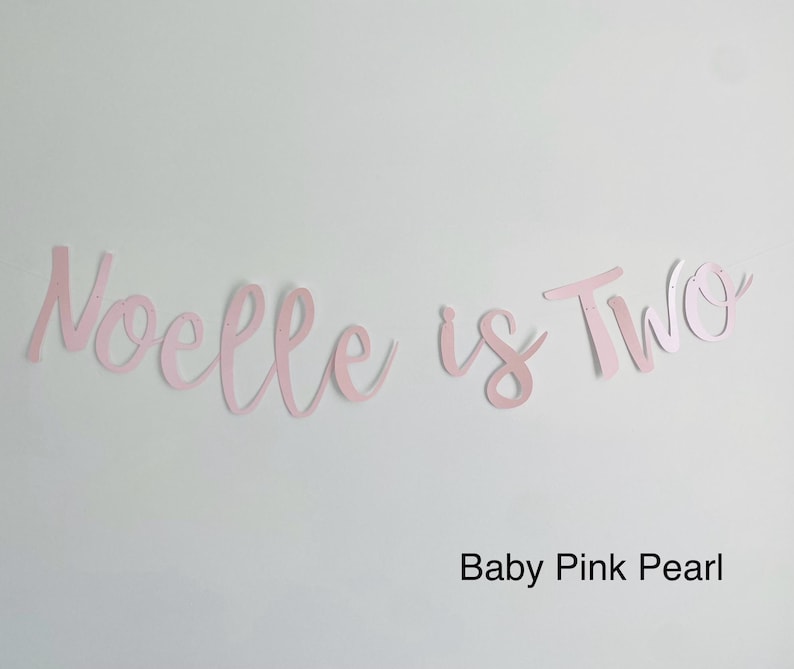 May include: Pink paper banner with the text "Noelle is Two" in a cursive font. The banner is on a white background. Baby Pink Pearl.