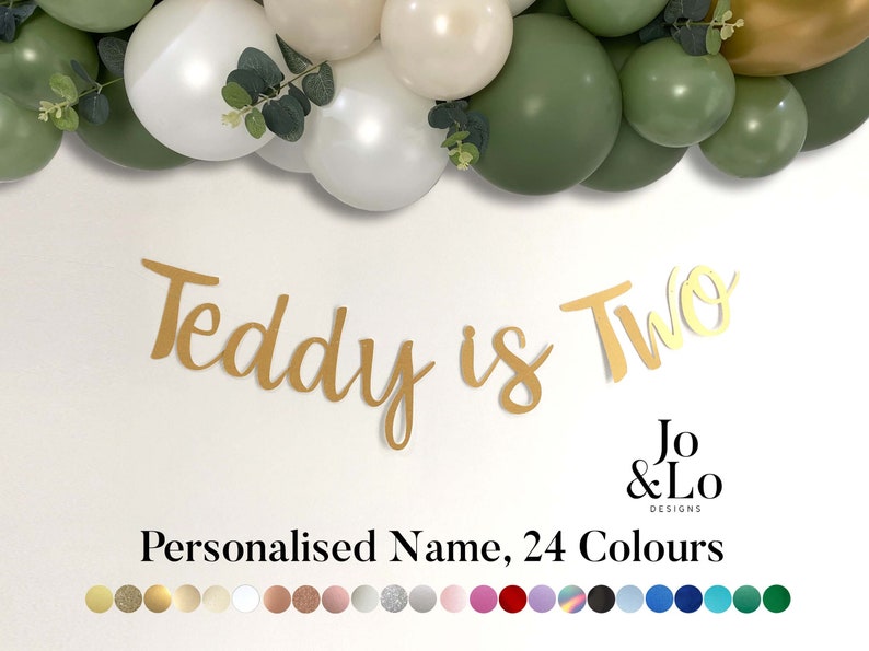 May include: Gold glitter banner with the text "Teddy is Two" and the logo "Jo & Lo Designs".  The banner is decorated with a garland of white and green balloons.  The banner is available in 24 colors.
