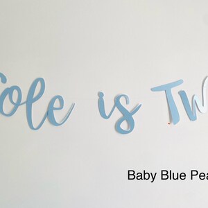 May include: A light blue banner with the text "Cole is Two" in a cursive font. The banner is made of paper and has a pearl finish. The text "Baby Blue Pearl" is written below the banner.