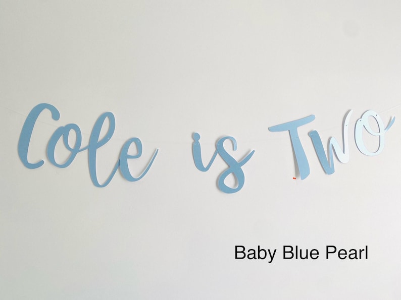 May include: A light blue banner with the text "Cole is Two" in a cursive font. The banner is made of paper and has a pearl finish. The text "Baby Blue Pearl" is written below the banner.