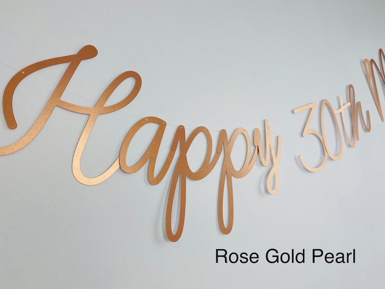 Pode incluir: Uma faixa de p&eacute;rola de ouro rosa com as palavras "Happy 30th" em fonte cursiva. A faixa &eacute; amarrada em uma corda fina e est&aacute; contra um fundo branco. O texto "Rose Gold Pearl" est&aacute; na parte inferior.
