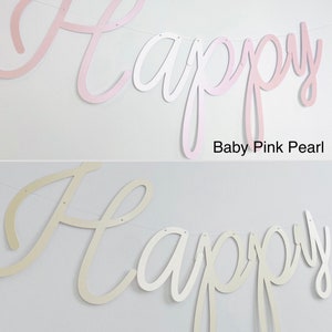 Pode incluir: Uma faixa com a palavra "Happy" em fonte cursiva, dispon&iacute;vel em Baby Pink Pearl e Ivory Pearl. As letras s&atilde;o unidas por uma fina corda branca. O fundo &eacute; uma parede branca lisa.