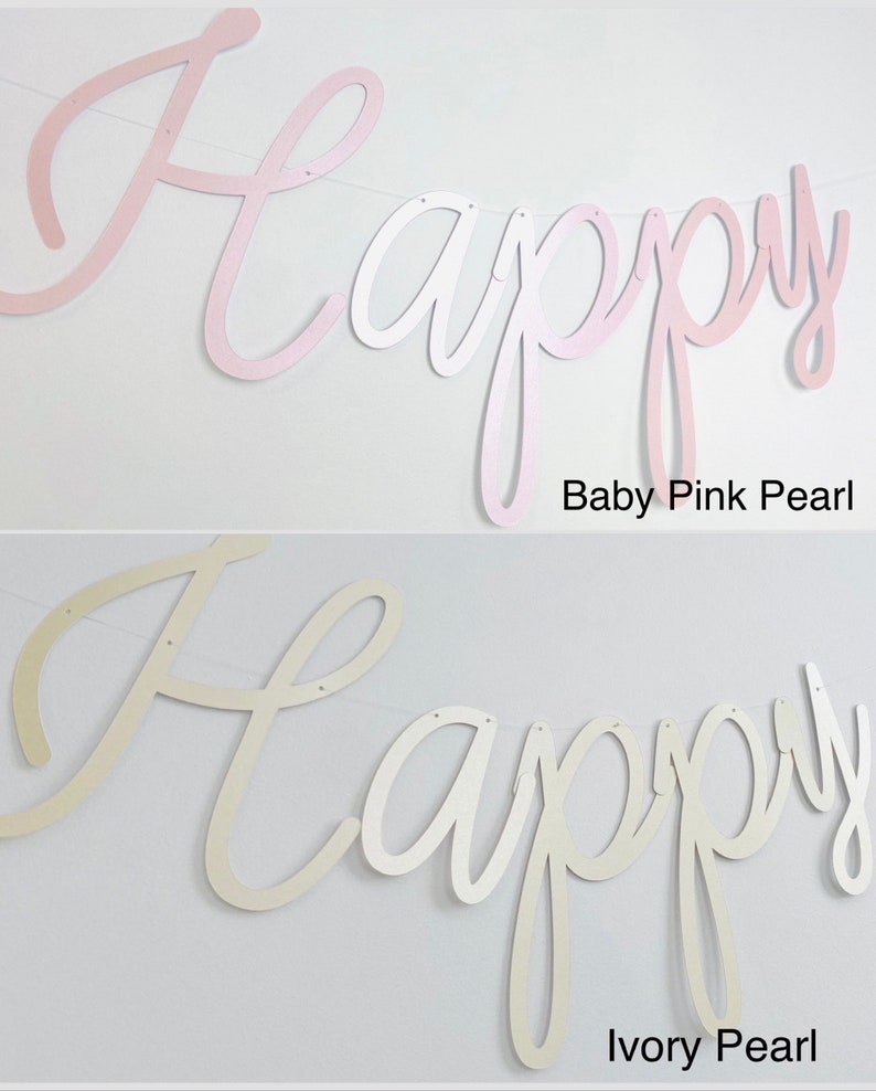 Pode incluir: Uma faixa com a palavra "Happy" em fonte cursiva, dispon&iacute;vel em Baby Pink Pearl e Ivory Pearl. As letras s&atilde;o unidas por uma fina corda branca. O fundo &eacute; uma parede branca lisa.