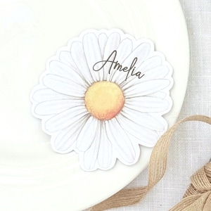 May include: A daisy-shaped sticker with the name "Amelia" in cursive script. The sticker has white petals and a yellow centre with a hint of orange. It rests on a white plate, with a brown ribbon in the background.