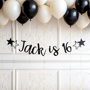 May include: A black script banner reading Jack is 16, flanked by black and white star cutouts, suspended by thin gold strings beneath a cluster of matte black and white balloons. The display features a cohesive monochromatic colour scheme.