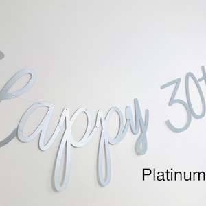 Pode incluir: Uma faixa prateada com as palavras "Happy 30th Jess" em fonte cursiva. A faixa &eacute; feita de um material met&aacute;lico e &eacute; amarrada em uma fina corda branca. As palavras "Platinum Pearl" est&atilde;o escritas abaixo.