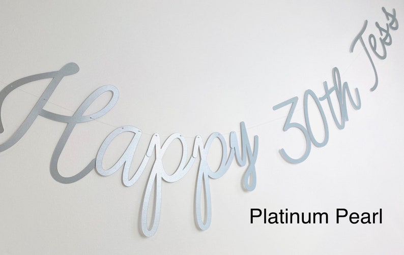 Pode incluir: Uma faixa prateada com as palavras "Happy 30th Jess" em fonte cursiva. A faixa &eacute; feita de um material met&aacute;lico e &eacute; amarrada em uma fina corda branca. As palavras "Platinum Pearl" est&atilde;o escritas abaixo.