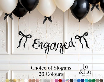 May include: An engagement party decoration featuring black and white balloons with black ribbon accents. A black banner spells out "Engaged" in cursive script. The image also includes text that reads "Choice of Slogans" and "26 Colours".