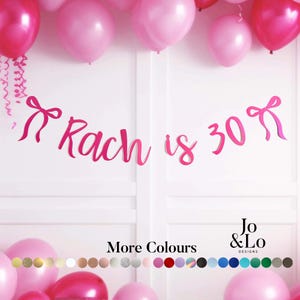 May include: A pink script banner reading Rach is 30 with a bow icon on each end. The banner is displayed against a white background with pink balloons at the top and bottom. The text More Colours is visible above a row of small circular colour swatches.