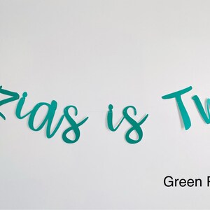 May include: A teal green banner with the text "Ozias is Two" in a cursive font. The banner is on a white background. Green Pearl.