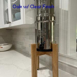 Op de afbeelding: Een waterfilter van roestvrij staal met een heldere afwerking, staand op een houten standaard. De standaard is gemaakt van eikenhout en heeft een natuurlijke afwerking. Het waterfilter staat op een aanrecht in een keuken.