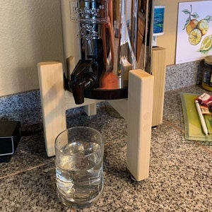 Op de afbeelding: Een waterdispenser van roestvrij staal met een houten standaard. De dispenser heeft een kraan en staat op een aanrecht. Een glas water staat voor de dispenser.