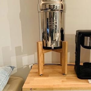 Op de afbeelding: Een waterfilter van roestvrij staal met een houten standaard. De filter staat op een houten aanrecht naast een zwarte koffiemachine.