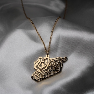 Peut inclure: Collier avec pendentif doré représentant le contour d'un pays, orné d'une calligraphie arabe complexe. Le pendentif est suspendu à une chaîne délicate, sur un fond de tissu blanc et soyeux.