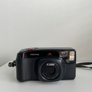 Peut inclure: Un appareil photo Pentax AF Zoom noir avec une sangle noire. L'appareil photo a un objectif argenté avec le texte "Pentax Lens 1:3.5/38mm-115mm" imprimé dessus. L'appareil photo a également un bouton rouge et le texte "AF Auto Focus" imprimé dessus.