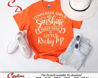 May include: Orange t-shirt with white text that reads "Tennessee Girls Are Sunshine Mixed With A Little Rocky Top". The shirt is laid out on a white surface with a straw hat and denim shorts.