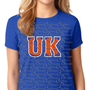 May include: A royal blue t-shirt with a white and orange basketball graphic and the letters "UK" in a blue outline. The shirt has a repeating pattern of the words "Carolina Designs" in a light gray color.