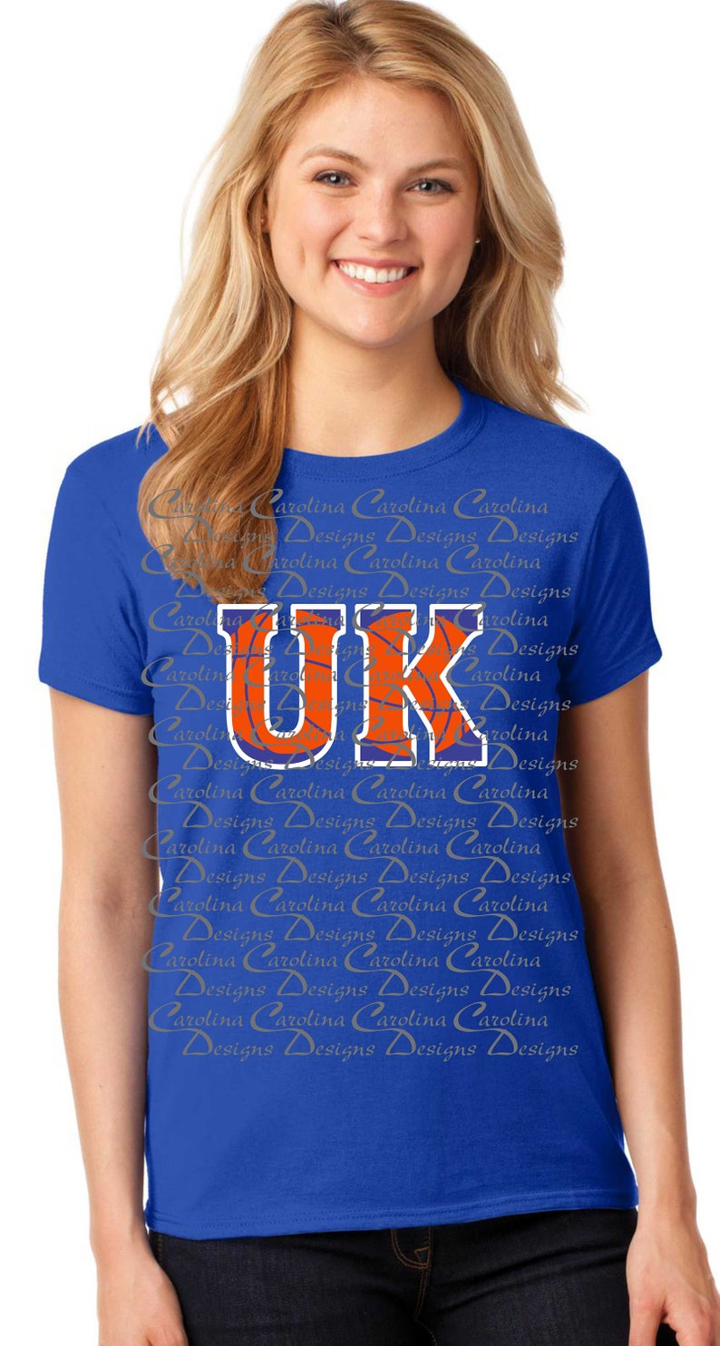 May include: A royal blue t-shirt with a white and orange basketball graphic and the letters "UK" in a blue outline. The shirt has a repeating pattern of the words "Carolina Designs" in a light gray color.