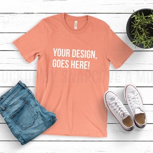 May include: A coral coloured t-shirt with the text "YOUR DESIGN, GOES HERE!" printed in white on the front. The t-shirt is laid flat on a white wooden surface with a pair of white trainers, blue jeans, and a potted plant.