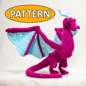 Könnte beinhalten: Ein gehäkelter rosa Drache mit hellblauen Flügeln. Der Drache hat einen langen Schwanz und steht auf einer weißen Oberfläche. Das Wort "PATTERN" ist in Orange auf einem gelben Oval geschrieben.