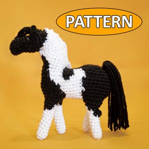 Op de afbeelding: Een gehaakt paardenspeelgoed in zwart en wit, met het woord "PATTERN" in een oranje ovaal erboven. Het paard heeft een zwarte manen en staart en witte benen. Het lichaam is een mix van zwart en wit haakwerk.