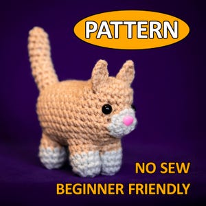 Könnte beinhalten: Gehäkelte Katze in Beige, Weiß und Rosa. Die Katze hat schwarze Augen und eine rosa Nase. Das Bild enthält das Wort "PATTERN" in einem gelben Oval und den Text "NO SEW BEGINNER FRIENDLY" in Weiß.